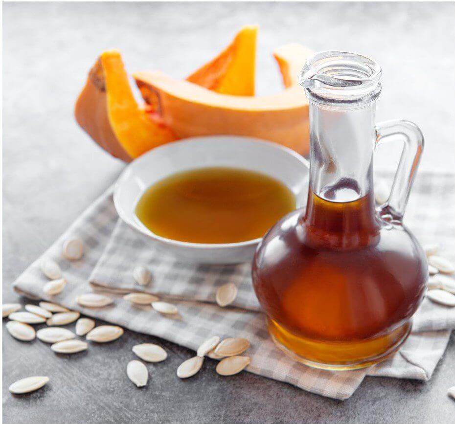 A glass bottle of liquid sits next to a bowl of oil, with slices of pumpkin and pumpkin seeds arranged on a checkered cloth.