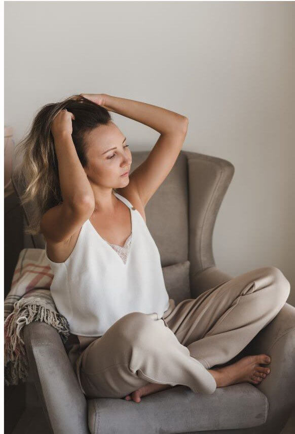 A woman with shoulder-length hair sits cross-legged in a chair while pulling her hair back with both hands.