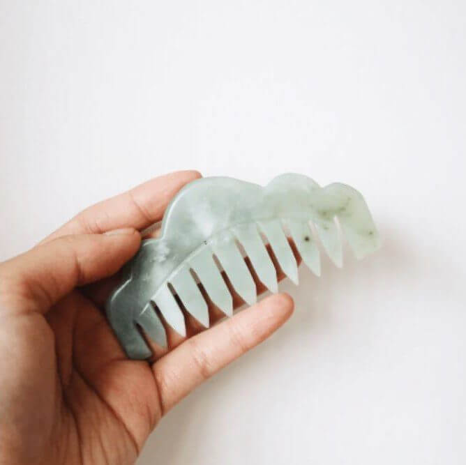 A hand holds a green jade hair comb against a light background.