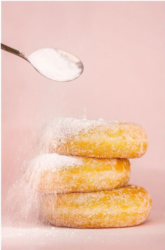 A stack of sugar-covered donuts beside a spoon on a white plate, showcasing a sweet treat ready to be enjoyed.