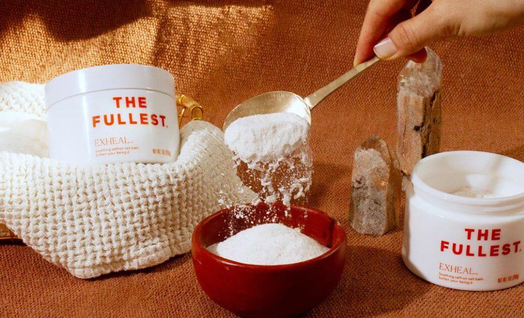A hand holds a spoonful of white powder above a red bowl while a container of the same powder sits beside it on a textured background.
