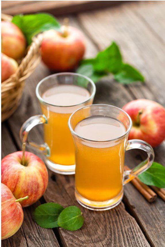 Two clear glasses of apple juice are situated on a wooden surface beside fresh apples and green leaves.