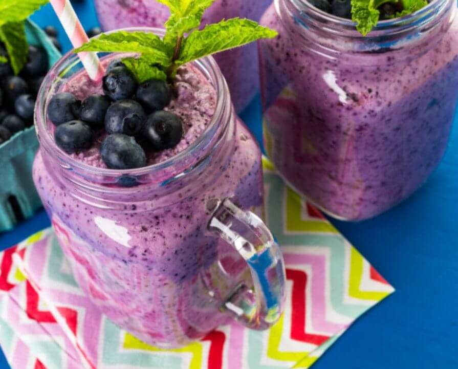 Frosted glass jars filled with berry smoothie are topped with blueberries and mint leaves, placed on a colorful zigzag patterned napkin against a blue background.