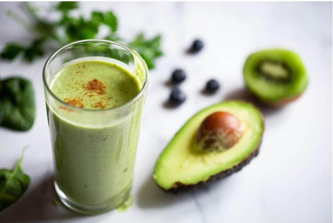 A tall glass of green smoothie garnished with a sprinkle of spice is placed beside halved avocado and blueberries on a marble surface.