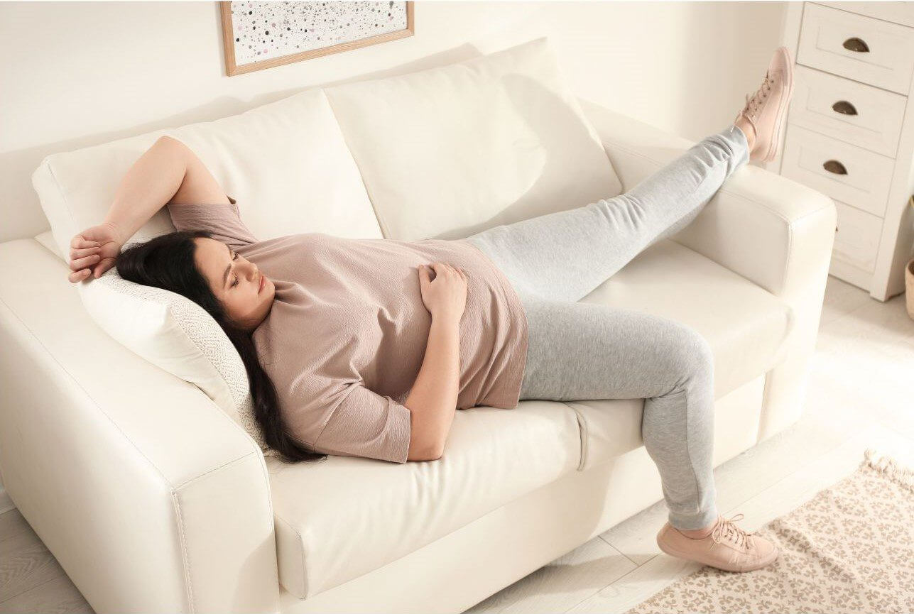 A person lies on a white couch with one arm resting on a pillow and the opposite leg extended outward while wearing gray leggings and a loose beige shirt.