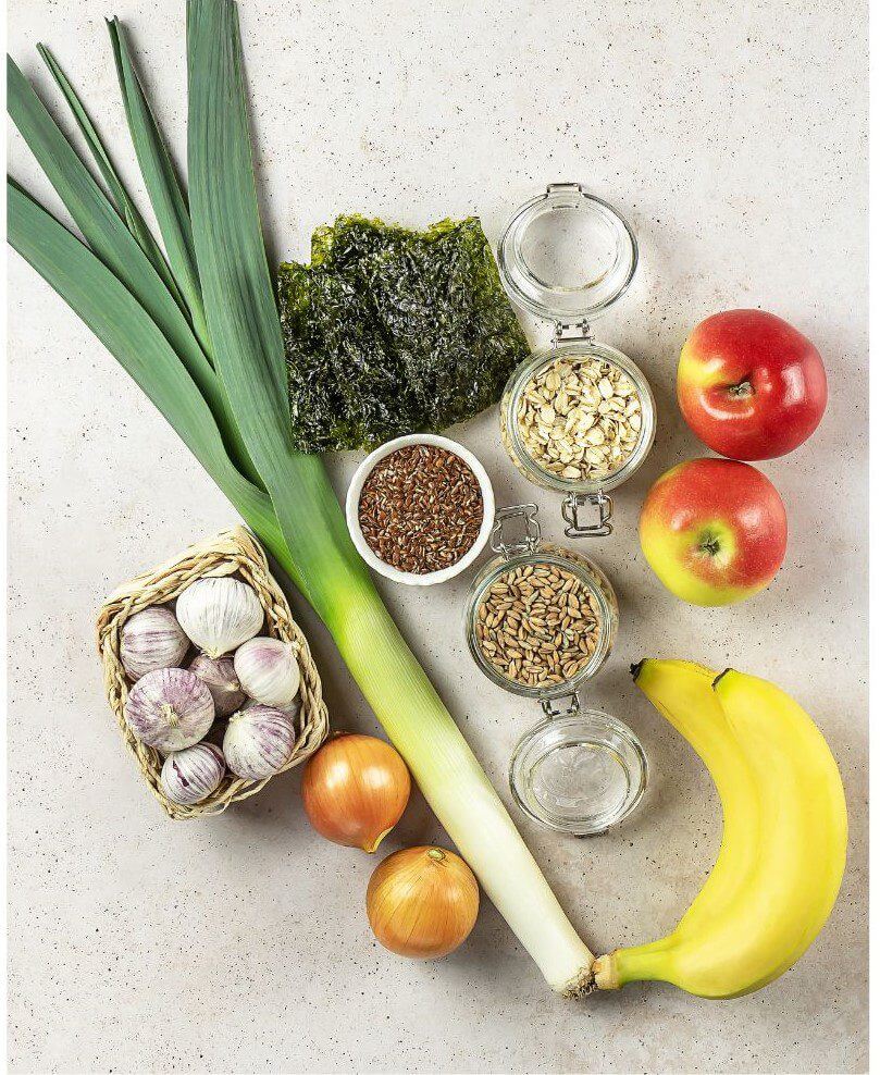 A variety of fresh fruits and vegetables, including leeks, apples, garlic, onions, and a banana, are arranged alongside jars containing seeds and oats on a light surface.