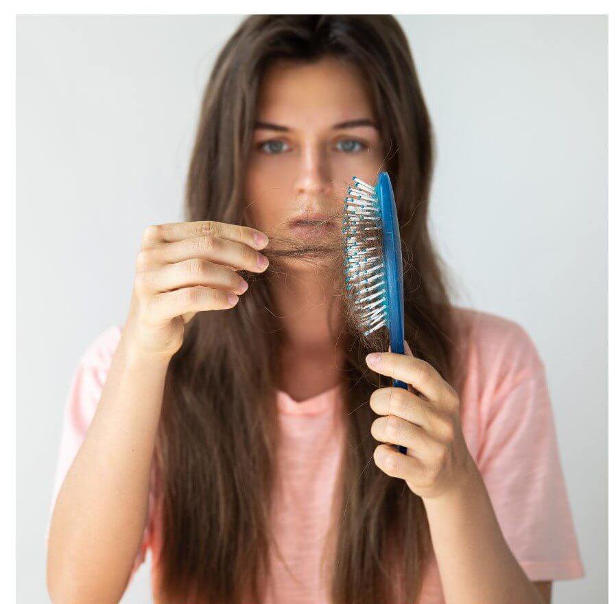 A woman in a pink shirt holds a strand of hair in one hand and a blue hairbrush in the other, looking at the camera with a concerned expression.