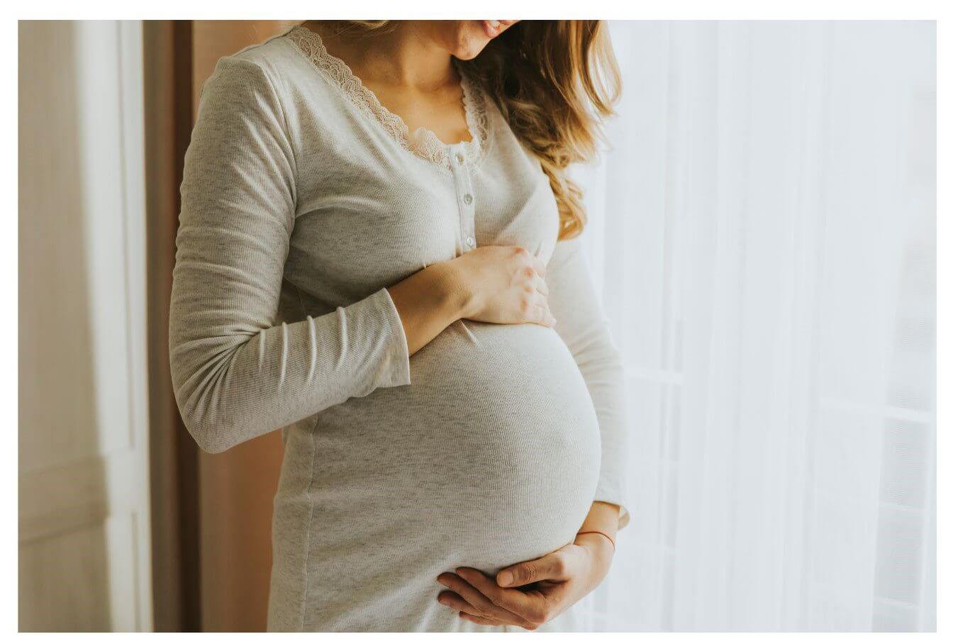 Pregnant woman in a cream-colored long-sleeved top cradling her belly with both hands while standing near a window.
