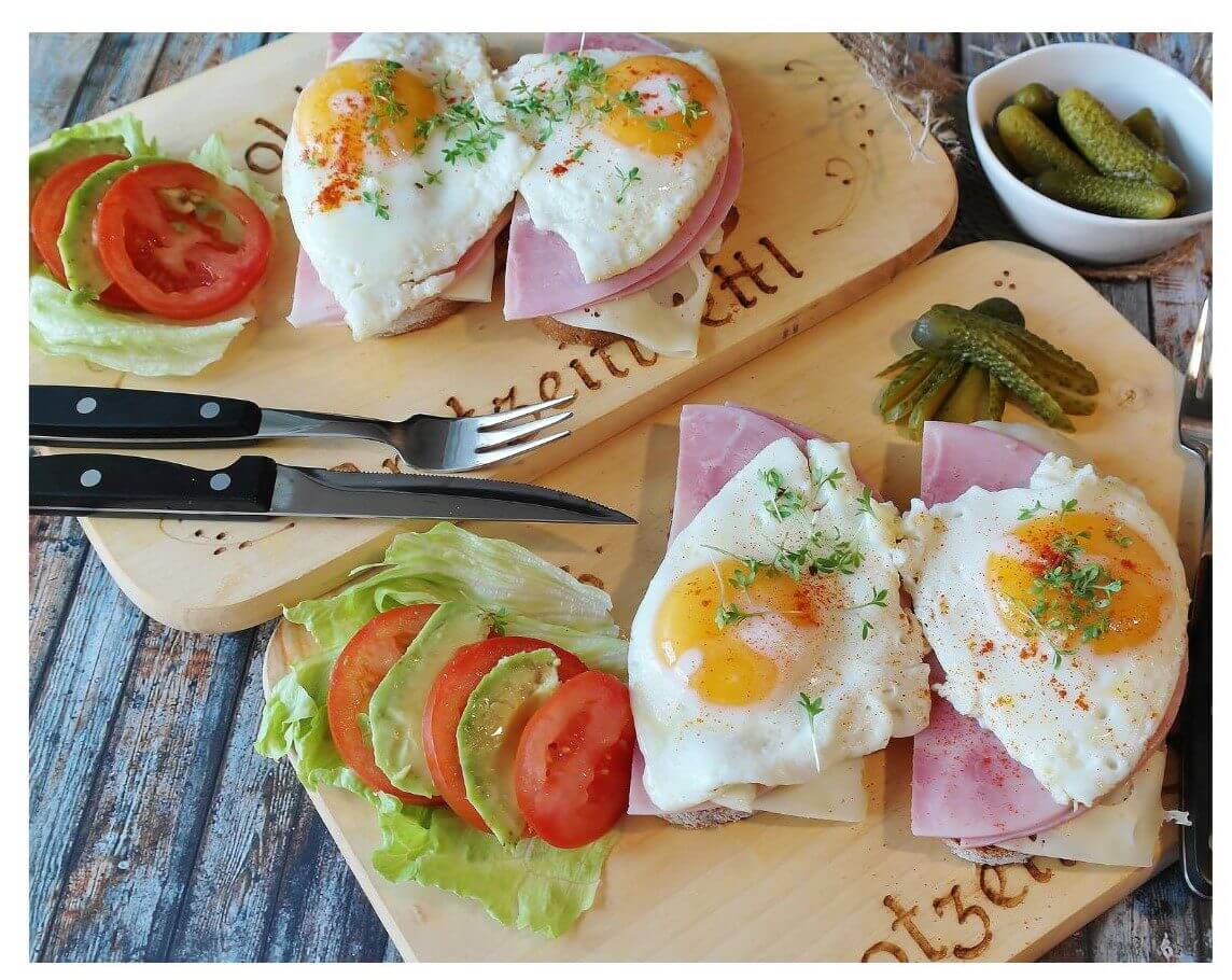 Two plates of open-faced sandwiches topped with fried eggs, ham, and garnished with herbs, served with slices of tomato, lettuce, and pickles.