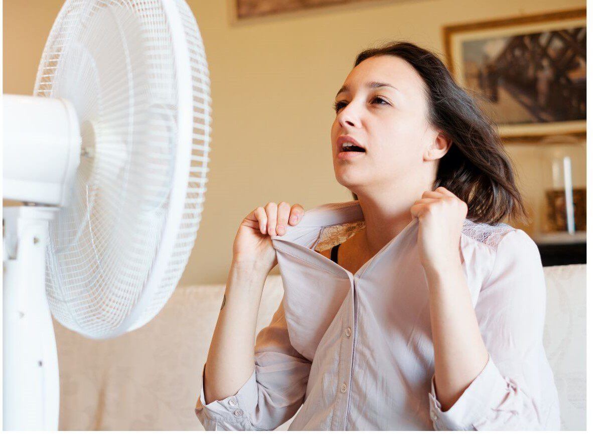 A woman is in front of a fan, pulling at her shirt while appearing to feel hot.