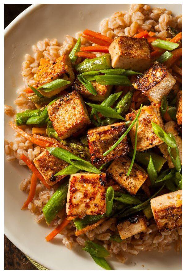 Stir-fried tofu cubes are arranged on a bed of farro with mixed vegetables including asparagus and carrots, topped with green onion slices.