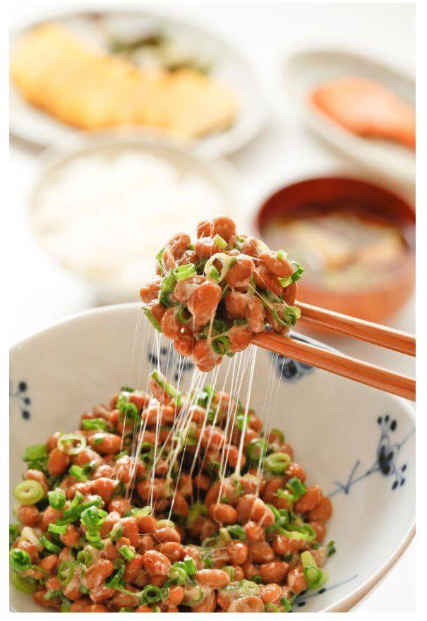 Chopsticks lifting a portion of diced salmon with green onions from a bowl as sauce drips down, with additional dishes blurred in the background.