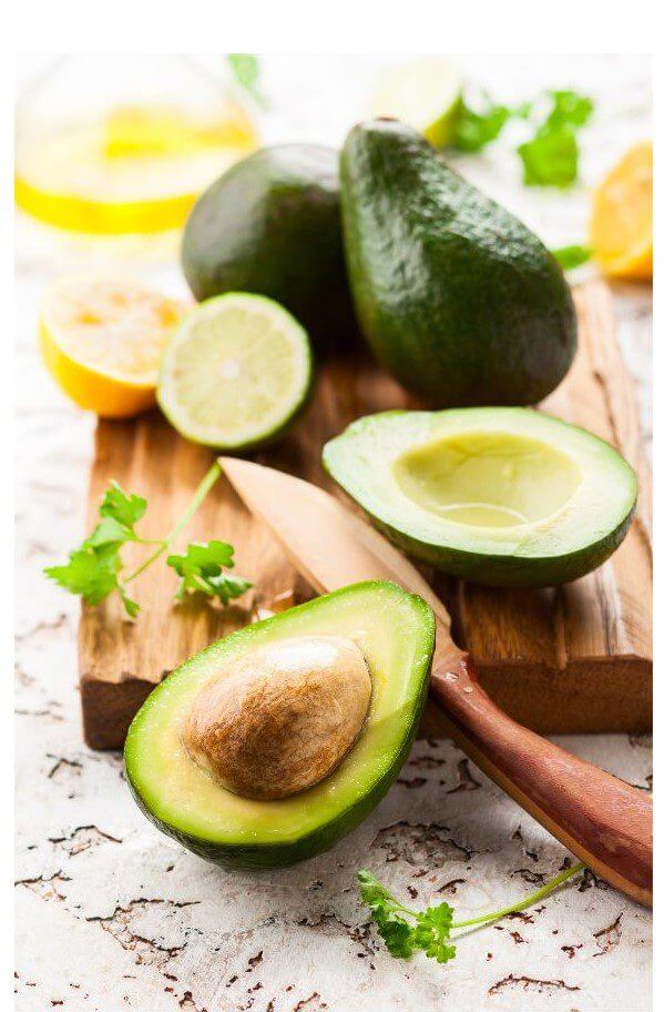 A halved avocado with a visible pit sits on a wooden cutting board alongside whole avocados and sliced citrus fruits.