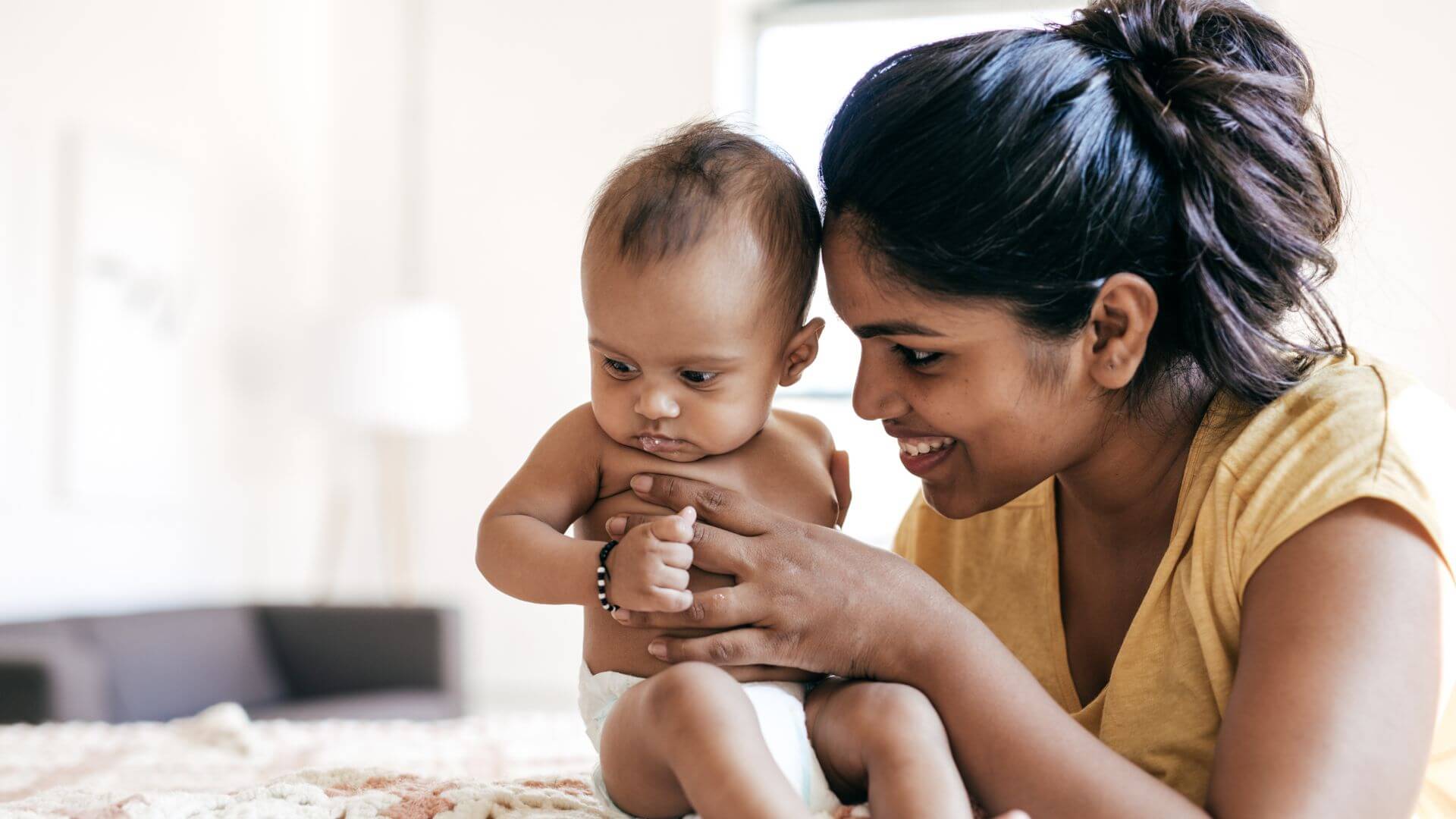 A woman in a yellow shirt is smiling while closely observing a bare-chested baby sitting in her lap.