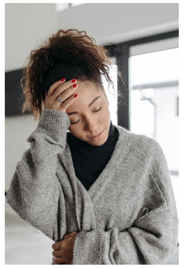 Woman with curly brown hair wearing a gray cardigan and black turtleneck holds her head with her hand while looking downward, appearing fatigued or experiencing discomfort.