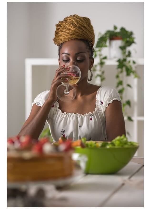 A woman with styled hair is drinking from a glass of wine while seated at a table with a salad and a cake in a bright indoor setting.