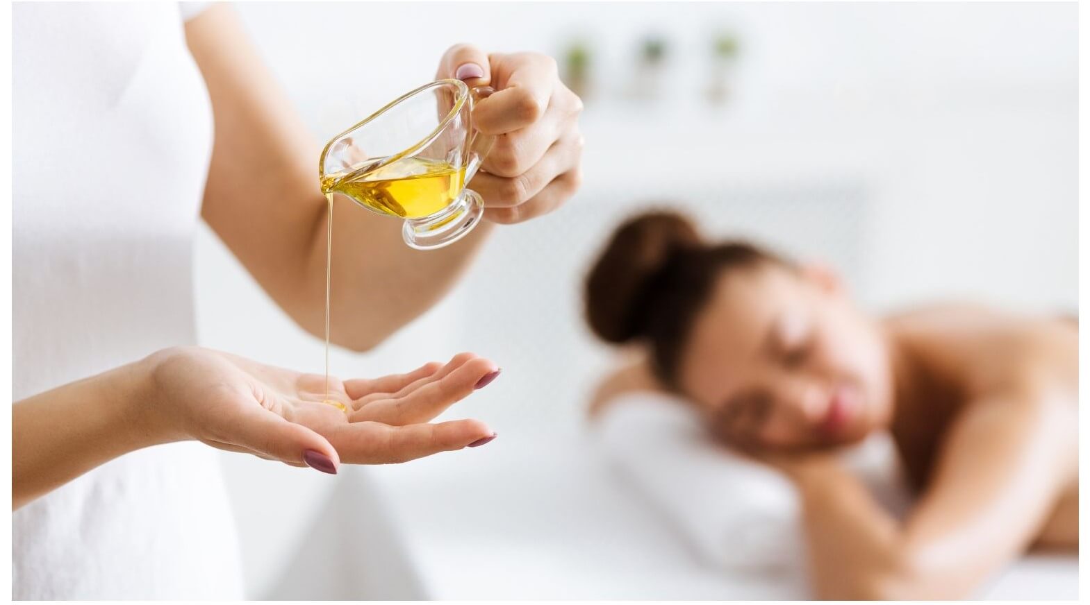 A person pours massage oil from a glass container into their hand while another person lies relaxed on a massage table in the background.