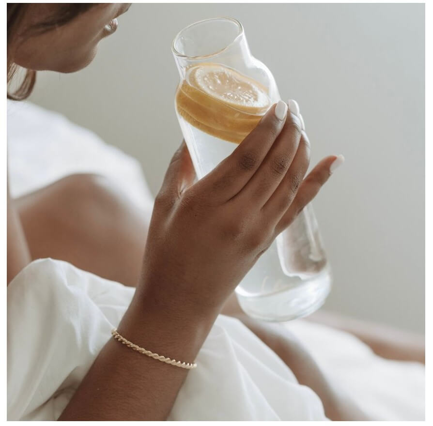 A person holds a glass pitcher of water with a lemon slice while resting on a bed.