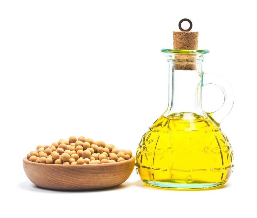 A glass oil jug filled with yellow oil stands beside a wooden bowl of beige soybeans.