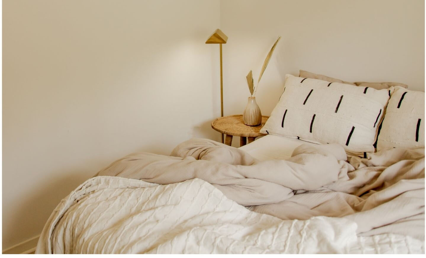 A neatly made bed with beige bedding and textured pillows is positioned next to a wooden side table featuring a decorative vase and plant.