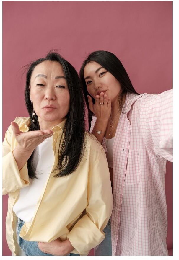 Two women pose for a selfie against a pink background, with one blowing a kiss and the other playfully touching her face.