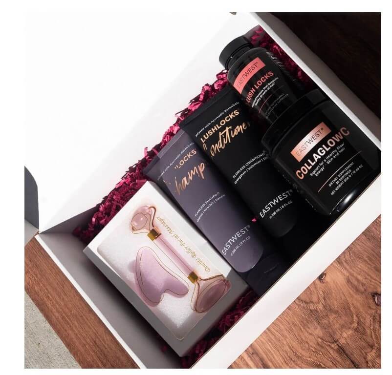 A white gift box contains hair care products and a facial tool, resting on a wooden surface.