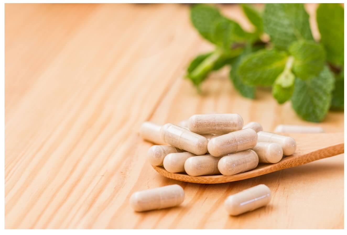 A wooden spoon holds a pile of beige capsules near fresh green mint leaves on a wooden surface.