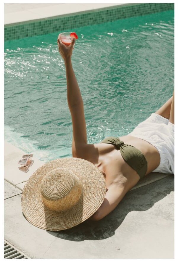 A person in a green bikini and straw hat lounges by a pool while holding a drink with one hand.