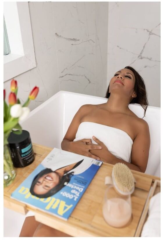 A woman wrapped in a white towel relaxes with her eyes closed in a bathtub, while a wooden tray holds a magazine and a skincare product nearby.