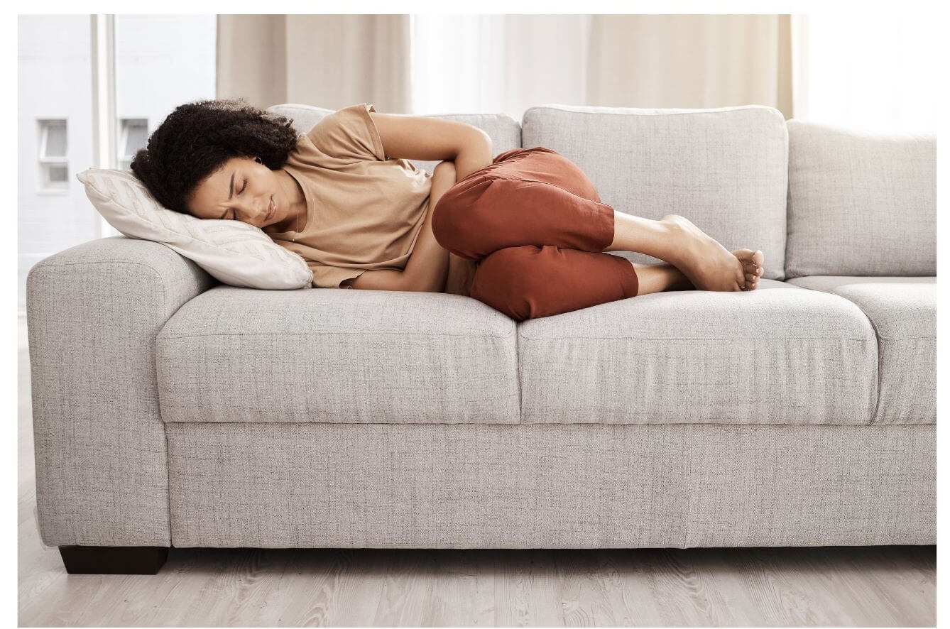 A person lies curled up on a light grey sofa with a pained expression, resting their head on a white pillow.