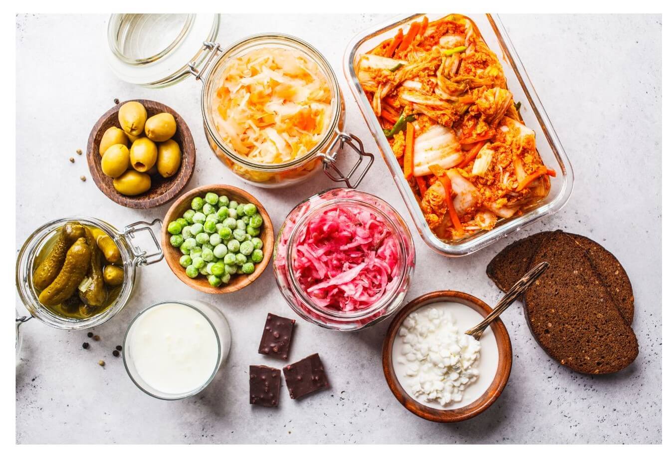 An assortment of fermented foods including jars of kimchi, sauerkraut, and pickles, along with bowls of cottage cheese, yogurt, dark chocolate pieces, and green peas, arranged on a light surface.