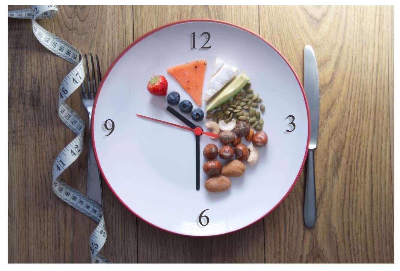 A clock-shaped plate arranged with various foods, including fruits and nuts, placed on a wooden surface beside a fork and knife.