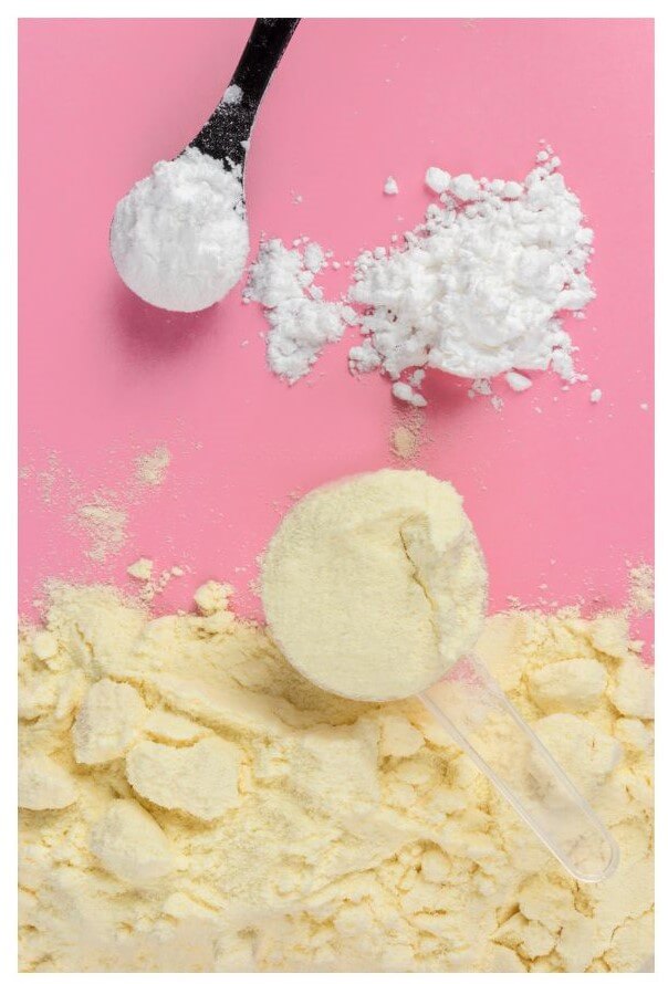 A black spoon holds white powder above a pile of yellow powder and a transparent scoop on a pink surface.
