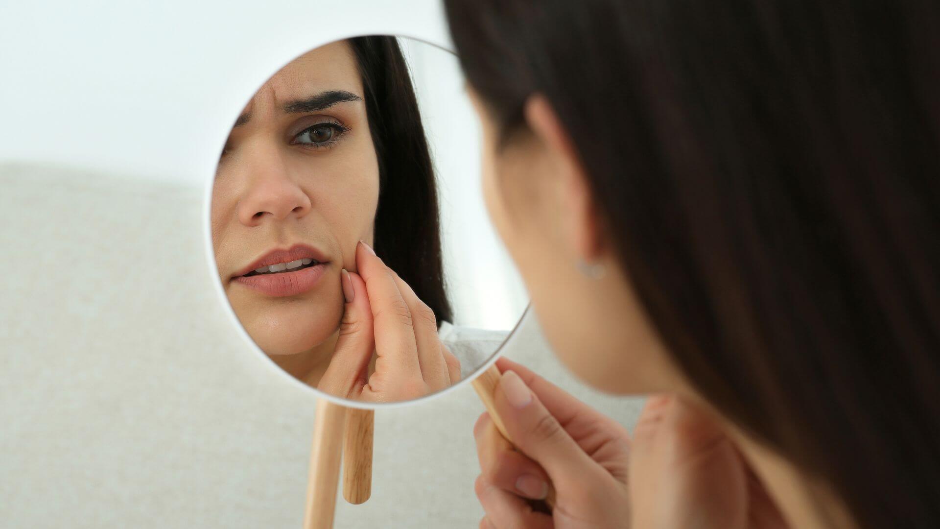A woman is closely examining her face in a mirror, touching her cheek with a concerned expression.