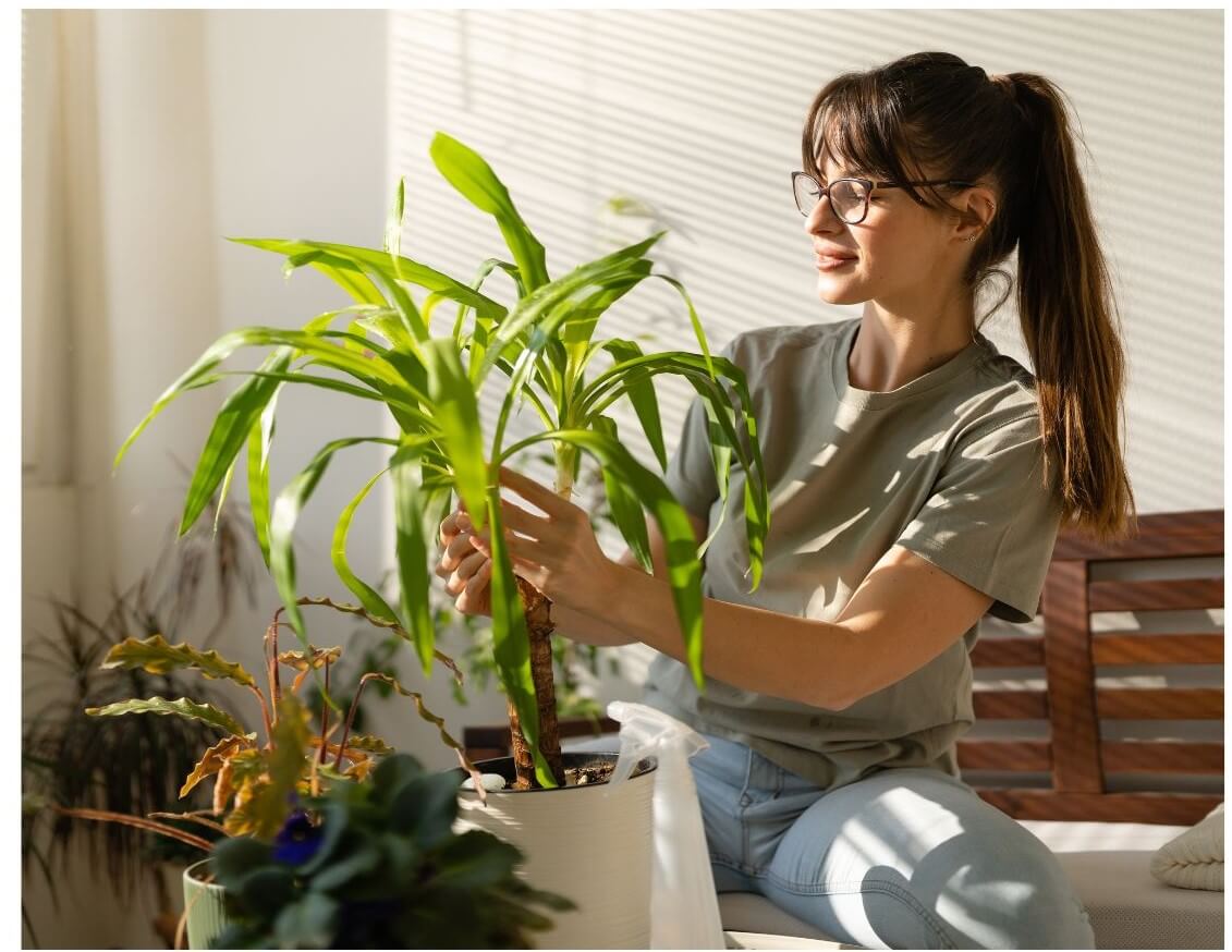 A woman with long hair and glasses is adjusting a potted plant while sitting on a couch in a well-lit interior space.