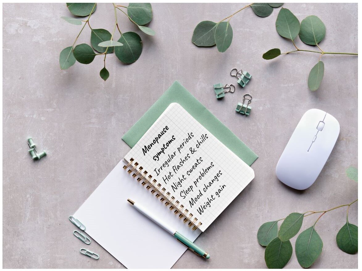 A notepad with handwritten notes about menopause symptoms rests on a gray surface alongside a pen, a computer mouse, green paper clips, and eucalyptus leaves.