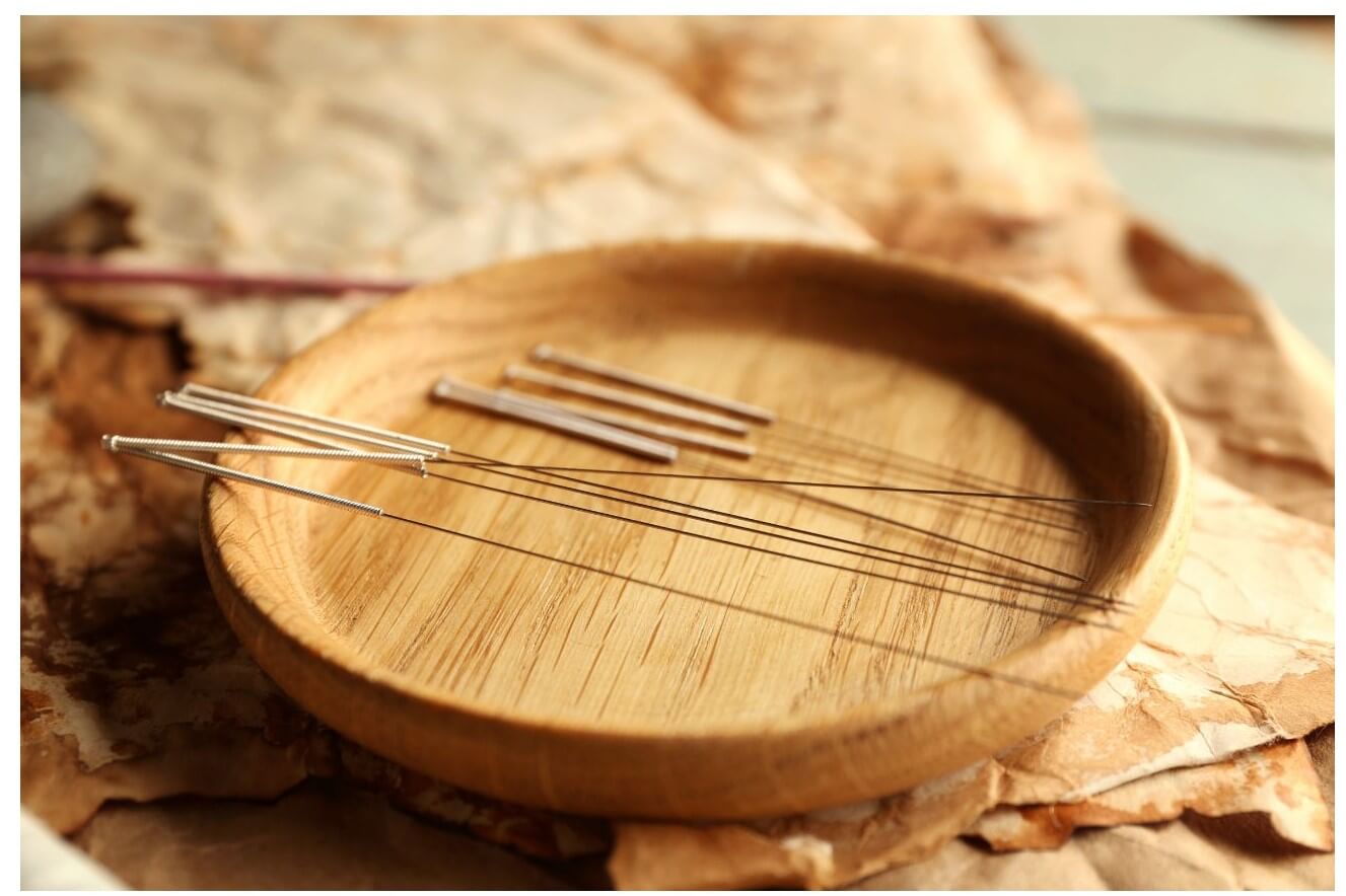 A wooden bowl contains several sewing needles arranged with their eyes facing upwards on a textured surface.