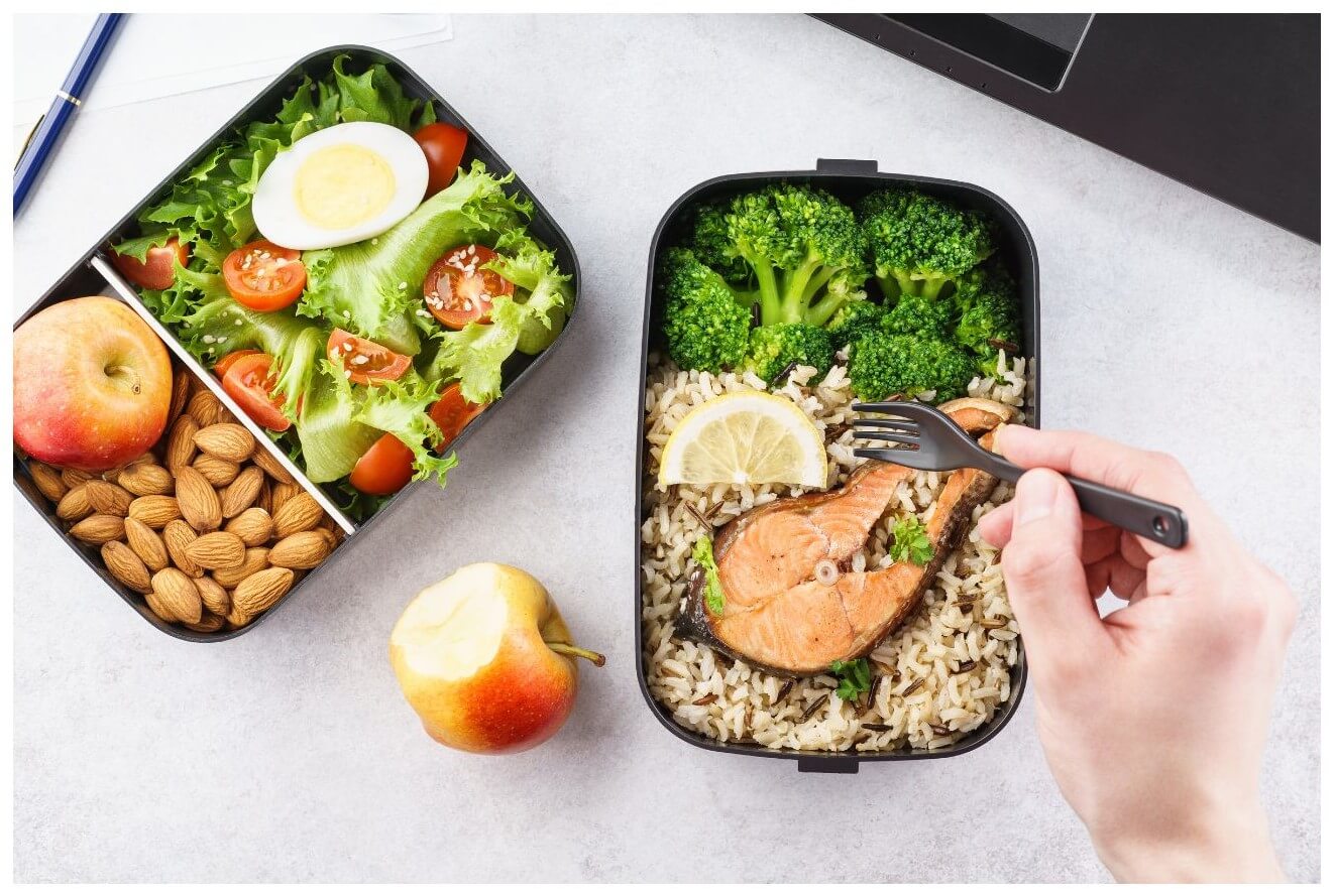 A hand holds a fork reaching for a salmon fillet on a bed of rice, accompanied by broccoli and a lemon slice, inside a divided lunch box.