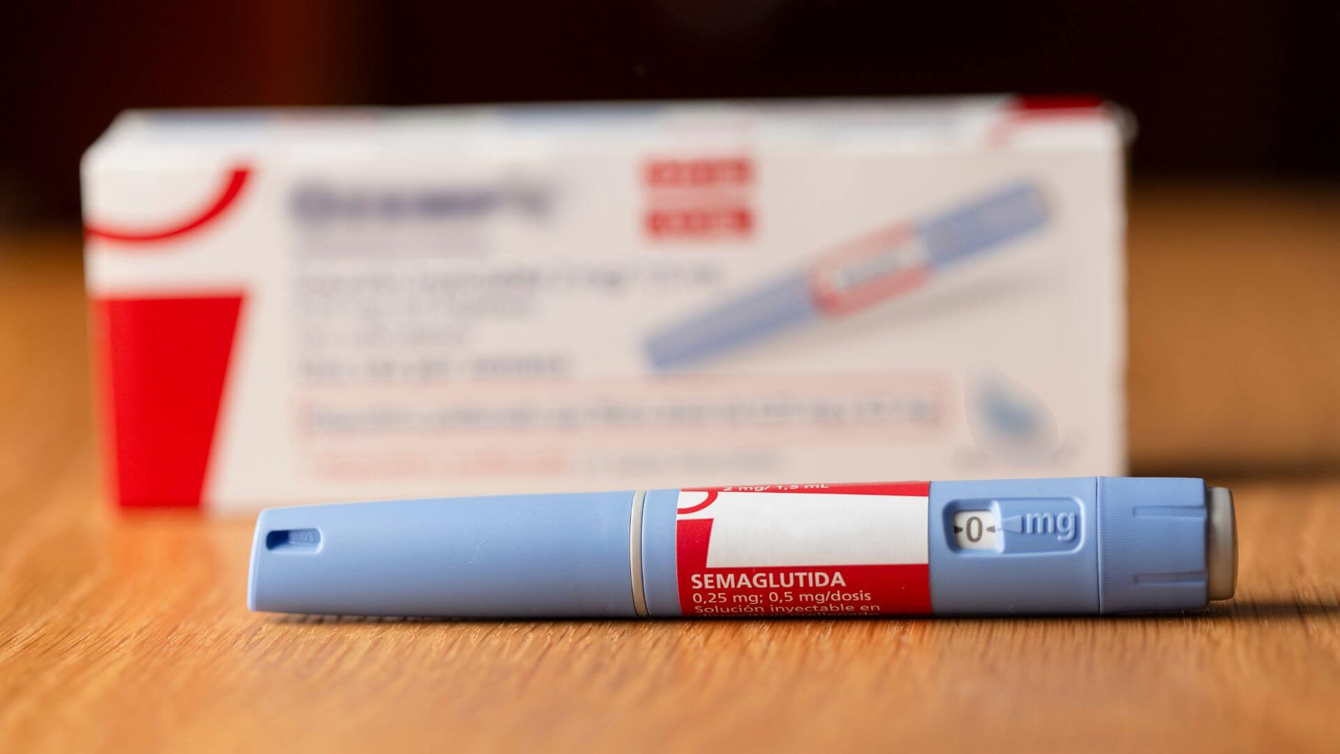 A blue and red medication injector is placed on a wooden surface, with a partially visible box in the background.
