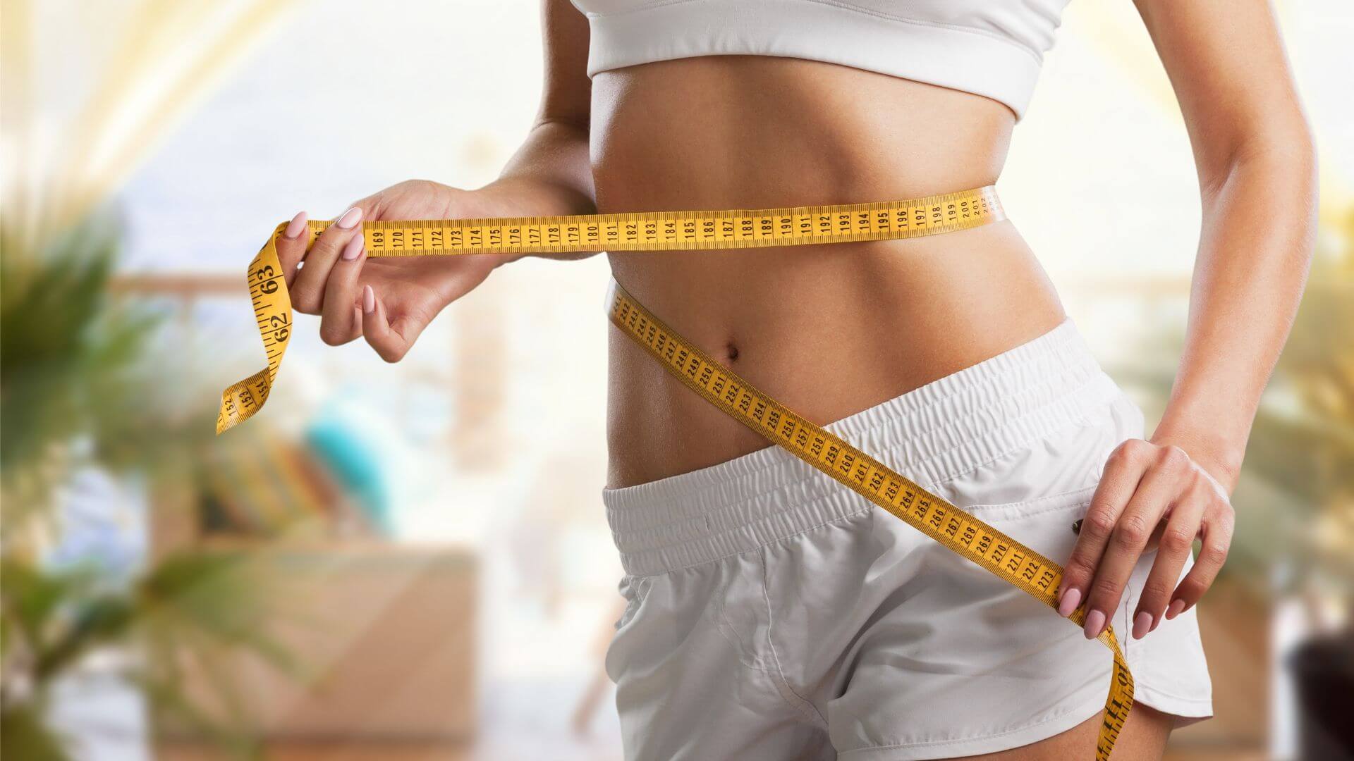 A person wearing white athletic shorts and a crop top is measuring their waist with a yellow measuring tape in a bright indoor setting.
