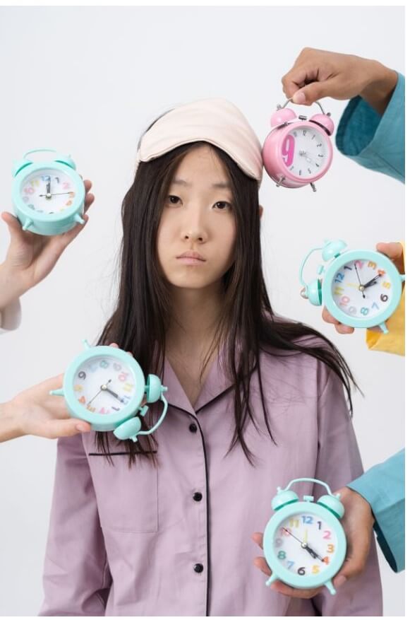 A young woman wearing a sleep mask looks tired while surrounded by multiple colorful alarm clocks being held by different hands.