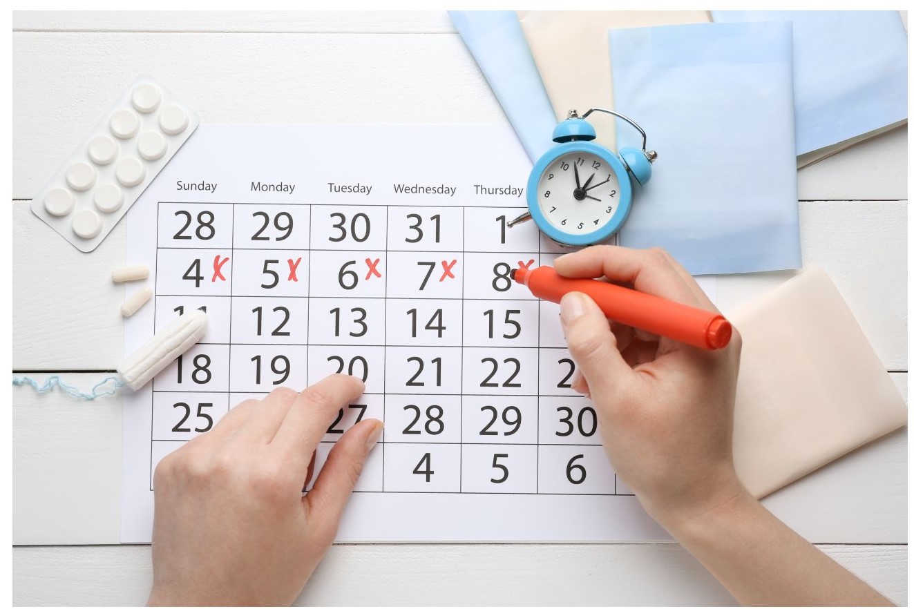 A hand with a red marker is marking dates on a calendar placed on a white wooden surface, surrounded by a blue alarm clock and small white pills.