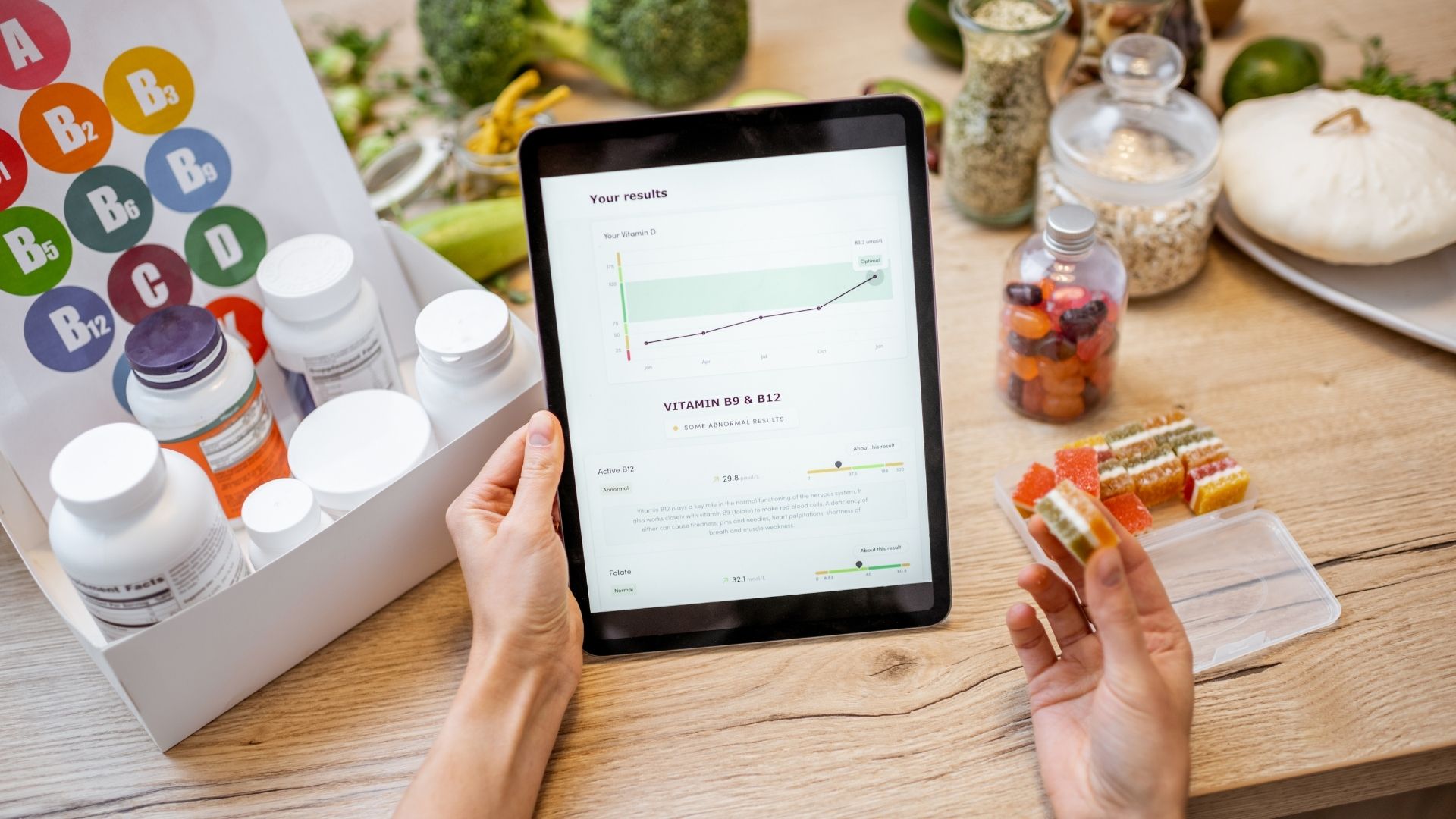 Person holding a tablet displaying vitamin D and B9 B12 test results with a graph, surrounded by supplement bottles and fresh produce on a wooden surface.