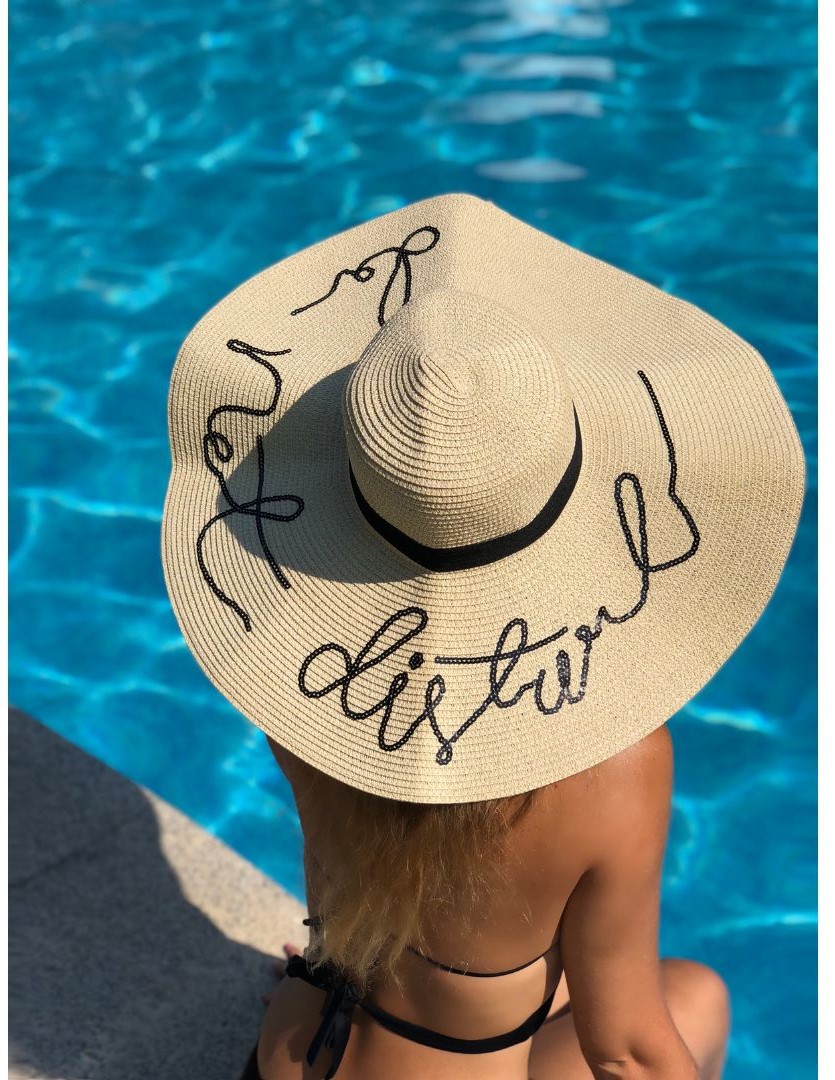 A person with long hair, wearing a black bikini, sits by a swimming pool while facing away, with a large straw hat that has the word 