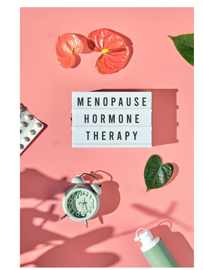 A flatlay composition on a pink background featuring a lightbox reading "MENOPAUSE HORMONE THERAPY" surrounded by birth control pills, a white alarm clock, red flowers, green leaves, and a cosmetic jar.