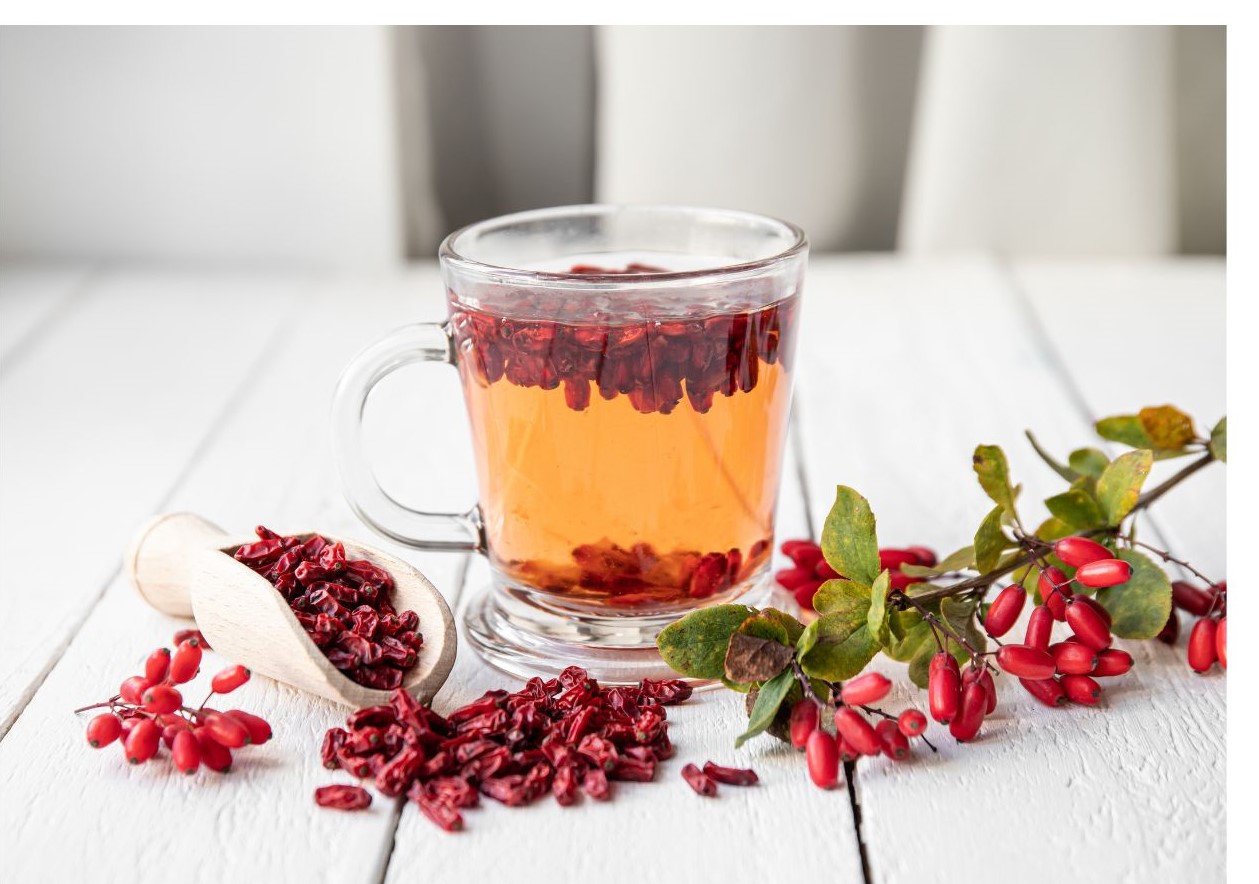 A glass cup filled with amber liquid containing red goji berries is placed beside a wooden scoop and sprigs of red berries on a white wooden surface.