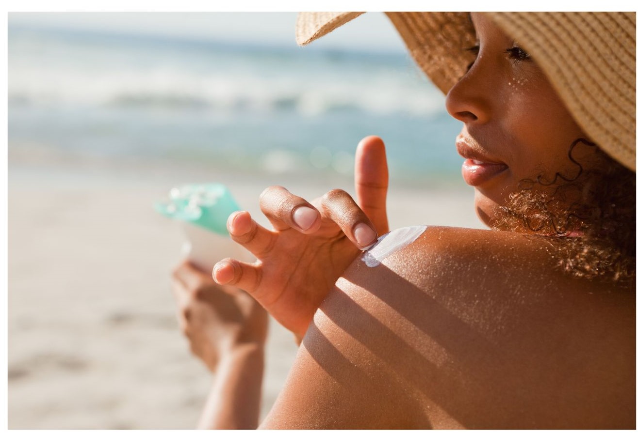A person applies sunscreen to their shoulder while sitting on the beach.