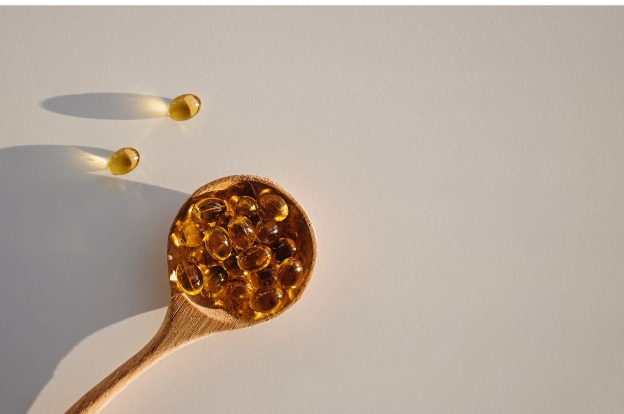 A wooden spoon holds several golden gel capsules, with two additional capsules placed nearby on a light-colored background.