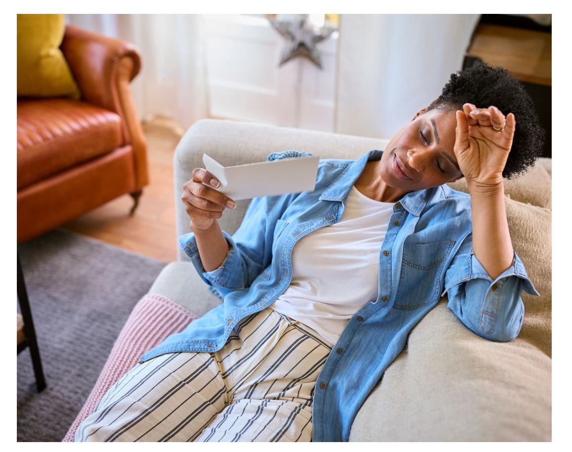 A woman with short, curly hair sits on a couch, looking at a letter while resting her forehead on her hand.