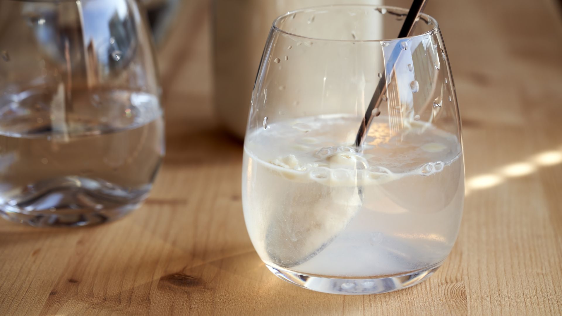 A glass of ice water with a black straw sits on a wooden surface alongside other glasses in soft natural light.